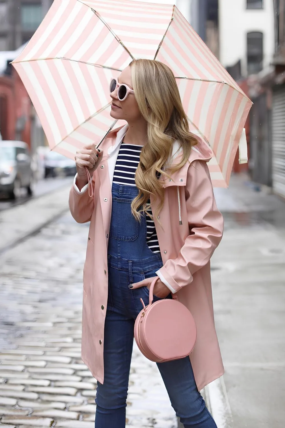 Woman wearing powder pink rain jacket paired with denim jumpsuit and a bag, topped with white and pink stripe umbrella